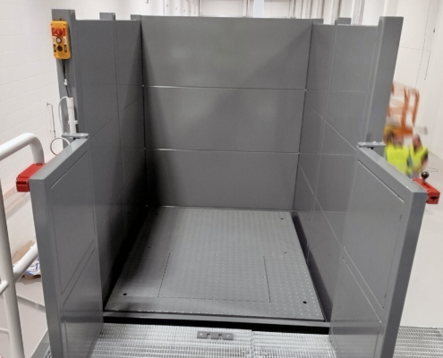 Offene Plattform des Hubtisches auf Übergabehöhe mit Einhausung und Sicherheitssteuerung Offene Einhausung eines Mehrfach-Scherenhubtisches mit geöffneter Plattform auf Hallenpodest. Die Plattform ist für den Transport von Gerätewagen ausgelegt.